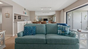 Un salon avec un canapé de couleur turquoise, deux coussins à motifs, une table basse blanche et, au fond, un coin cuisine.