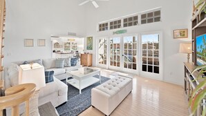 Ein großzügiges Wohnzimmer mit einem weißen Sofa, einem Glastisch und Holzboden. Ein großes Fenster bietet einen Ausblick nach draußen.