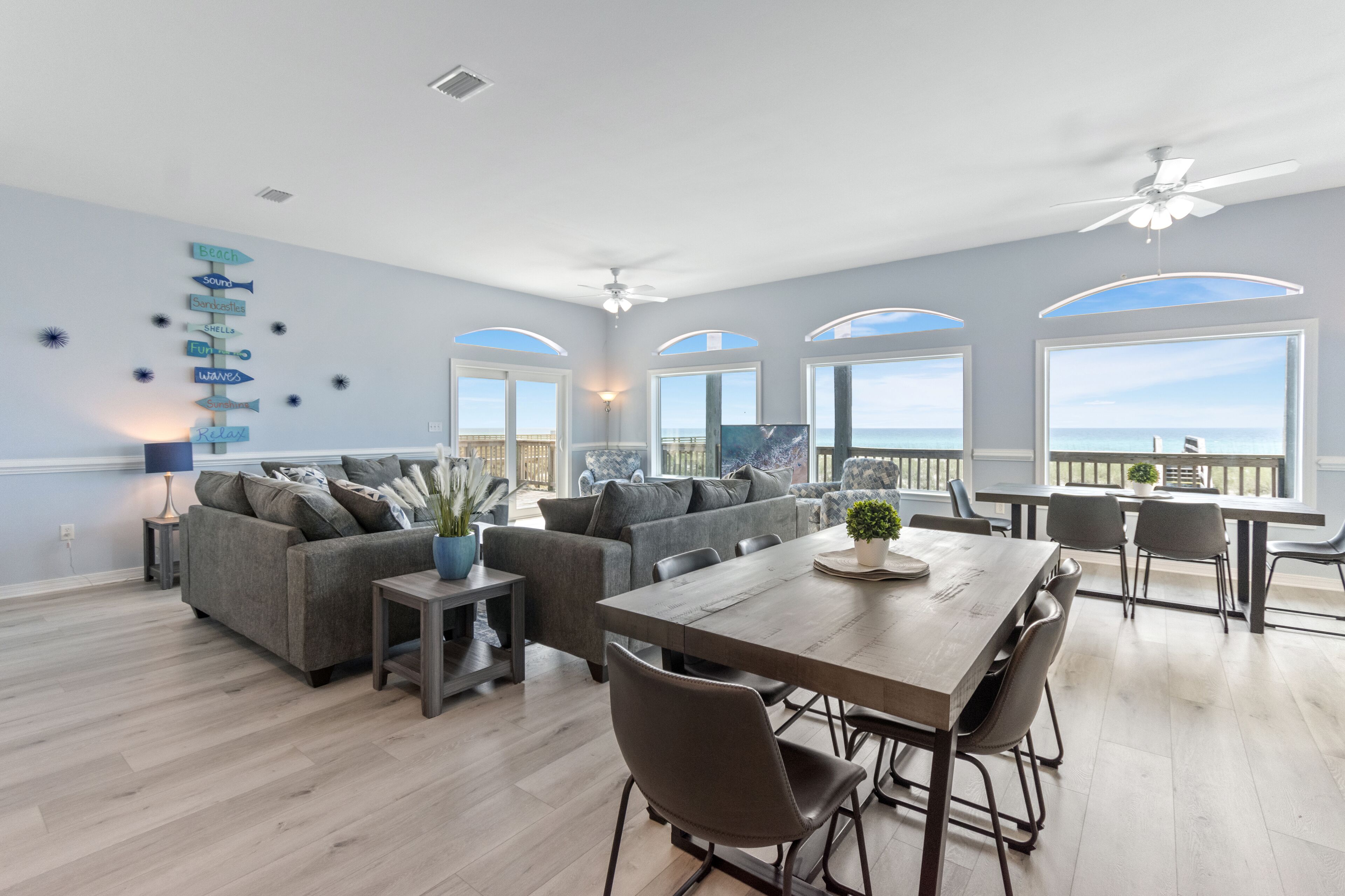 A spacious living room with a large dining table, grey sofas, and a view of the ocean through arched windows.