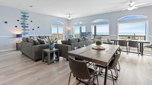 Ein geräumiges Wohnzimmer mit großem Esstisch, grauen Sofas und Blick auf das Meer durch Bogenfenster.