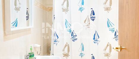 Une salle de bain avec un lavabo blanc, un miroir et un rideau de douche orné de motifs de voiliers.