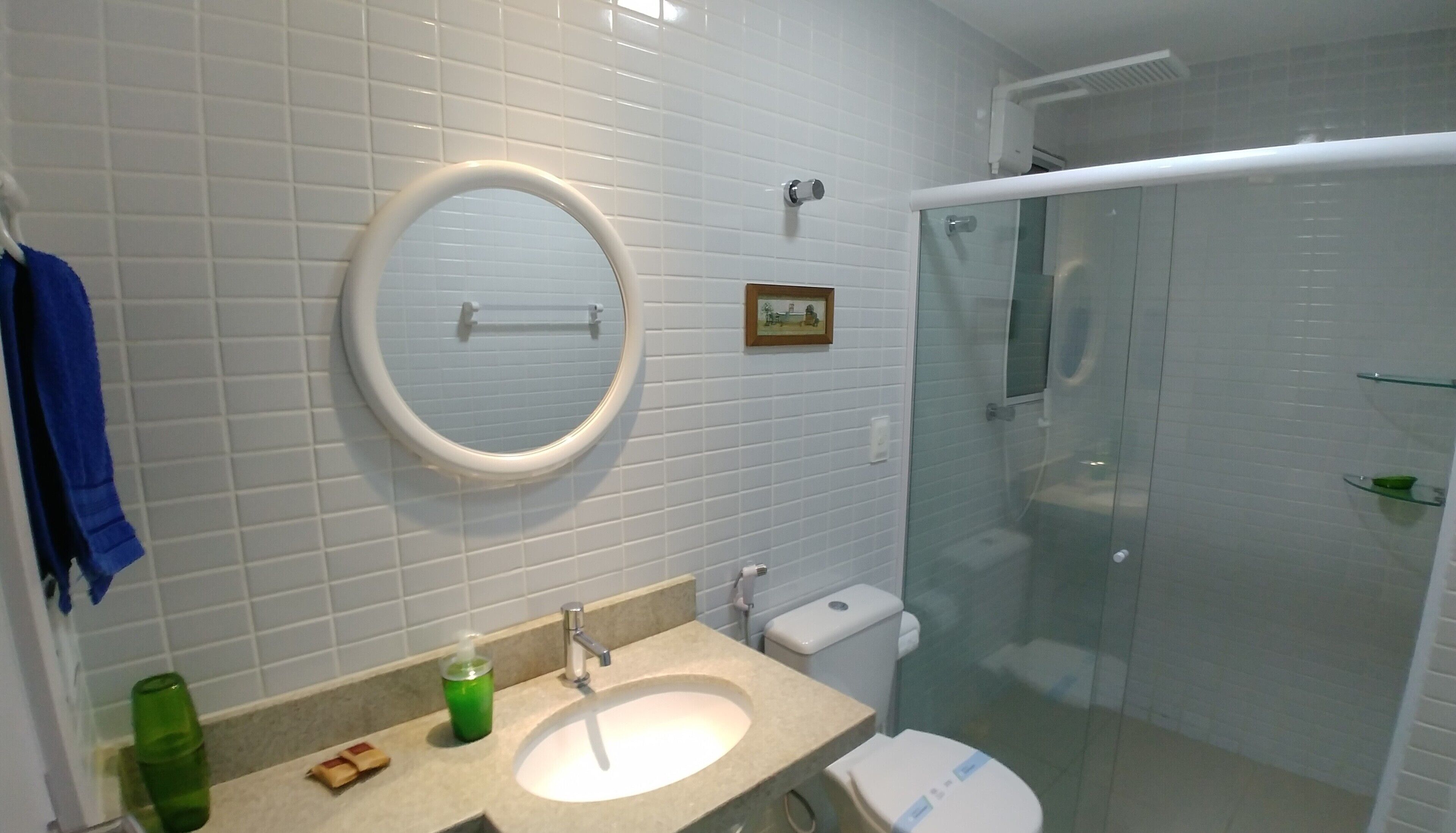 Une salle de bain avec des murs carrelés de carreaux blancs, un miroir rond, un lavabo avec une bouteille verte, des toilettes et une cabine de douche en verre.