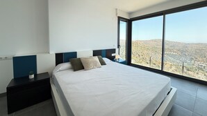 A modern bedroom with a large bed, a nightstand, and a view of a mountainous landscape through a large window. - Agi Canyelles Mar Casa 1 Casa for rent in Roses Girona. (Roses)