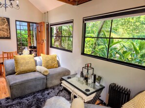 A modern living room with a grey sofa, two yellow cushions, a glass coffee table, and a view of lush greenery through large windows. - Cabin in the woods (Cape Town)