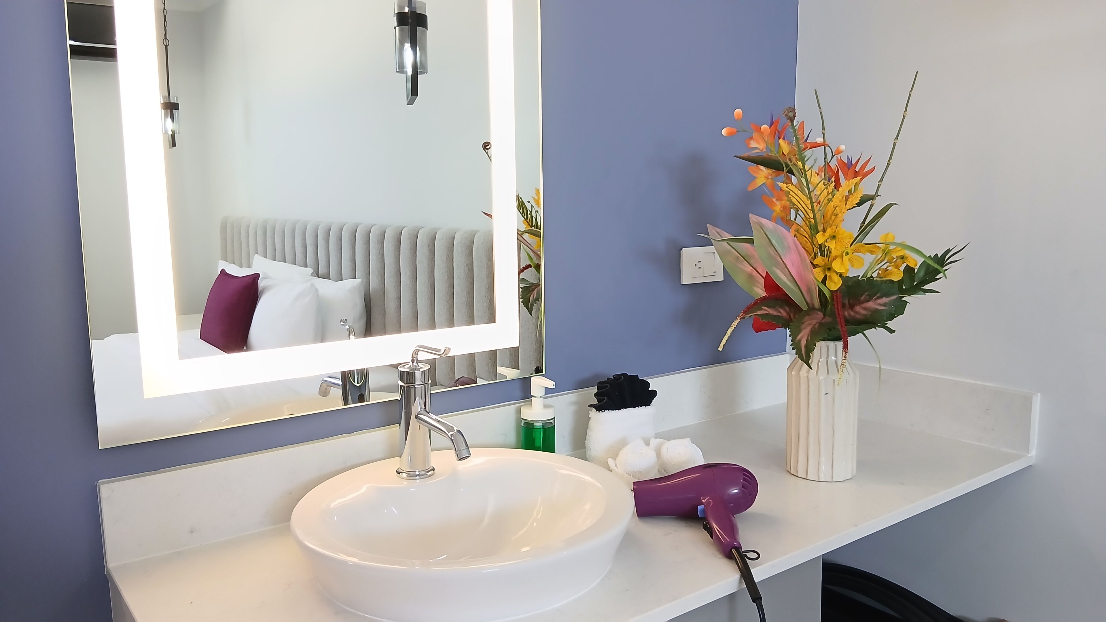A bathroom with a white sink, a mirror, a towel rack, a hairdryer, and a vase with flowers.