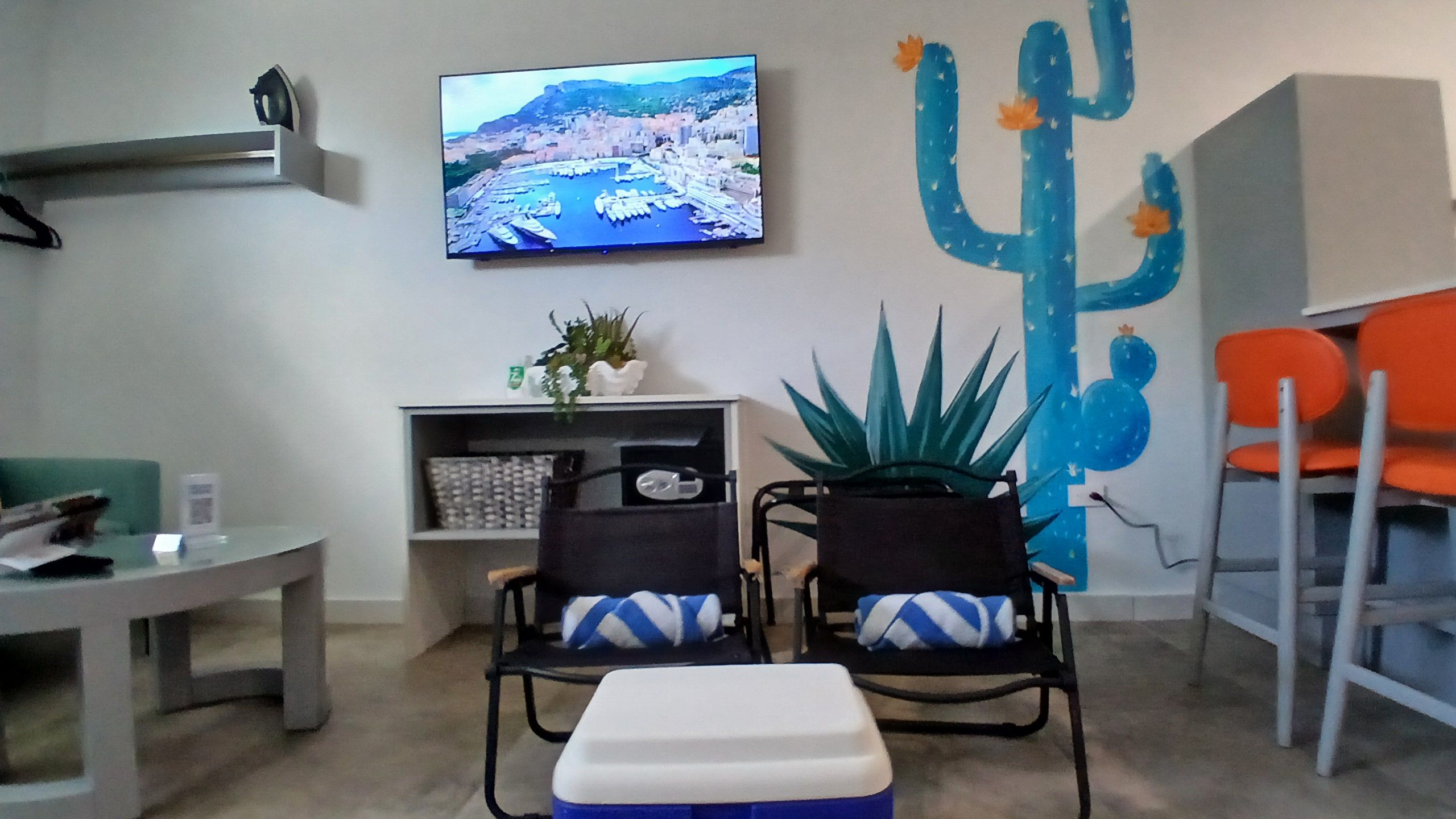 A room with a television mounted on the wall, a cactus decoration, two chairs with cushions, a small table, and a bench.