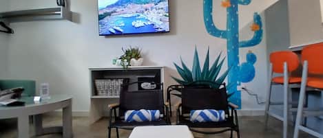 A room with a television mounted on the wall, a cactus decoration, two chairs with cushions, a small table, and a bench.