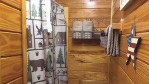 Une salle de bain rustique, avec une cabine en bois, un rideau de douche orné de motifs animaliers, une étagère avec des serviettes et un drapeau accroché au mur.