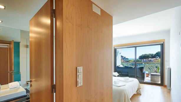 A hotel room with a wooden door, a bed with a white bedspread, a small table with a towel, and a view of the outdoors through a large window. - La Scala Sea Views - by Portugal Portfolio (Cascais)
