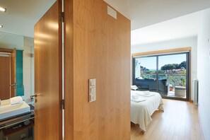A hotel room with a wooden door, a bed with a white bedspread, a small table with a towel, and a view of the outdoors through a large window. - La Scala Sea Views - by Portugal Portfolio (Cascais)
