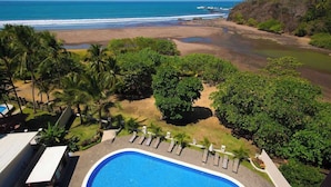 A pool with a view of the beach and ocean, surrounded by greenery and a building. - 7th Floor Ocean View. Sleeps 8. 3 Beds 3 Bath (Jacó)
