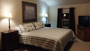 Une chambre à coucher avec un lit, deux tables de chevet avec des lampes, une télévision, une armoire, un tableau encadré au mur et une fenêtre avec des rideaux.