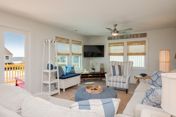 A spacious living room with a large window overlooking a beach, a ceiling fan, and a flat-screen TV mounted on the wall.