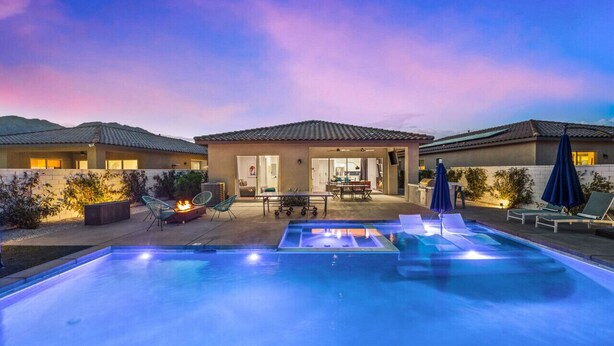 A swimming pool with lounge chairs and a fire pit, surrounded by a patio area with a view of a house.