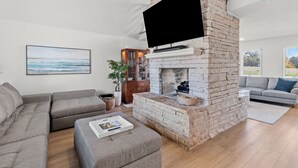 A living room with a brick fireplace, a flat-screen TV, grey sofas, and a large window. - Spacious KC Home - Pickleball, Mini Golf, Gym, Luxurious Bathrooms, Swim Spa (Bonner Springs)