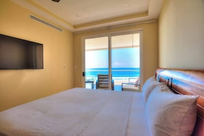 A bedroom with a large bed, a television mounted on the wall, and a sliding glass door leading to a balcony with a view of the ocean.
