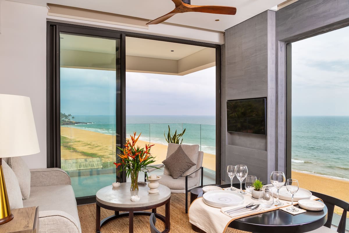 Una habitación moderna con balcón que da a una playa, amueblada con un sofá, una mesa de comedor para dos personas y un televisor en la pared.