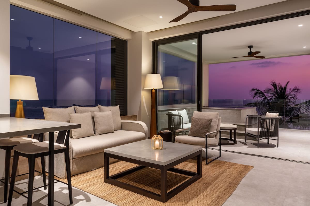 Una sala de estar moderna con sofá, mesa de centro y sillas, ventanas amplias con vistas, y ventiladores de techo.