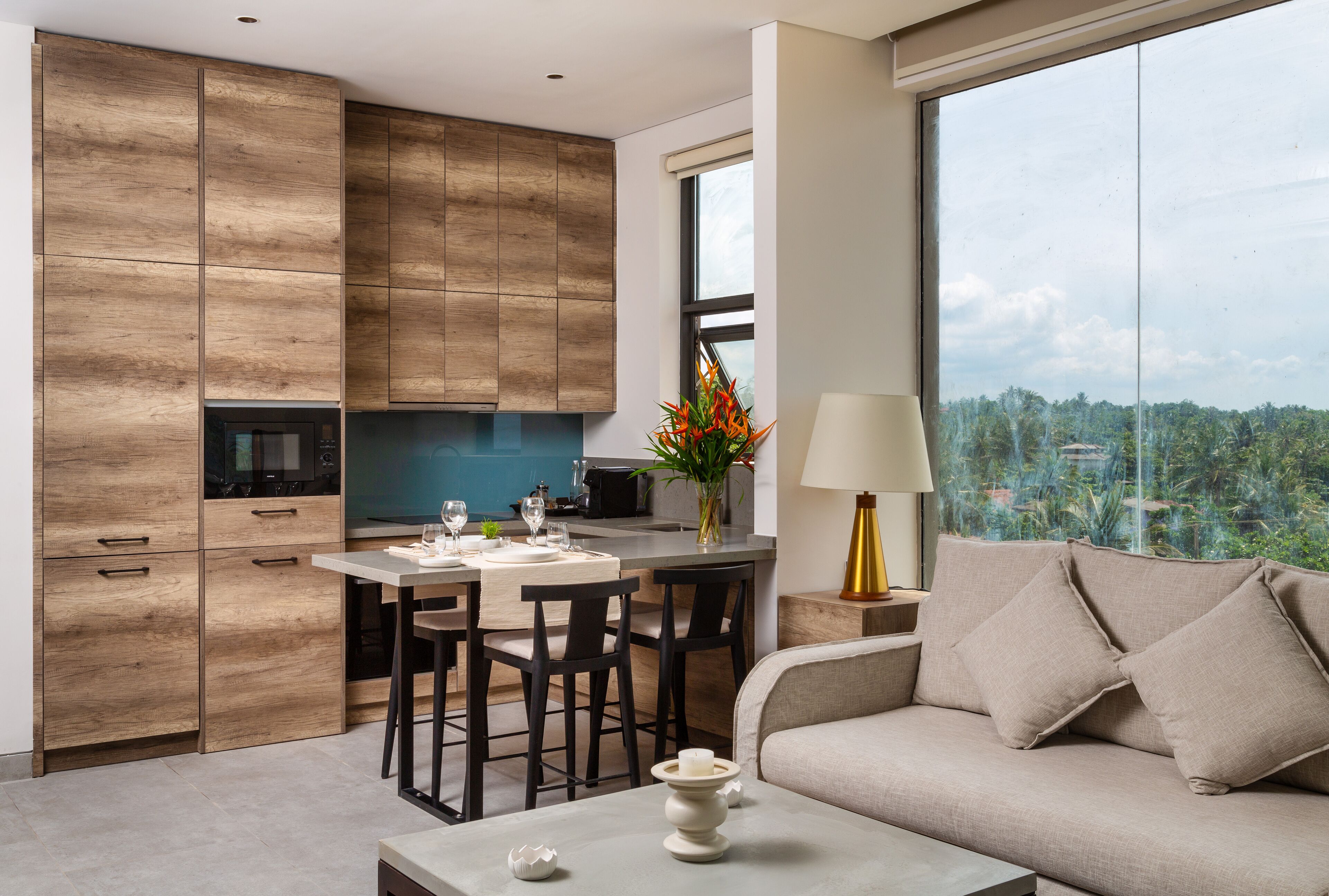 A modern kitchen with wooden cabinets, a dining area with a table and chairs, and a sofa with a view of greenery.