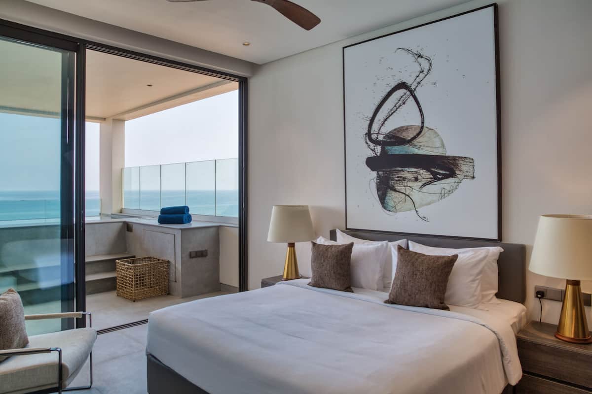 Una habitación de hotel moderna con una cama grande, dos lámparas de noche, un cuadro abstracto de gran tamaño, vistas al océano y un balcón con una cesta de mimbre.