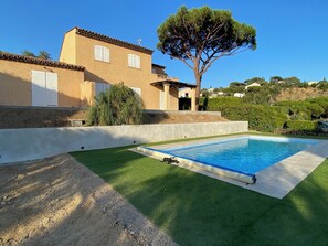 A swimming pool with a surrounding grassy area, a building with a tiled roof, and a large tree.