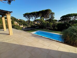A swimming pool with a tiled deck, surrounded by trees and a pergola.