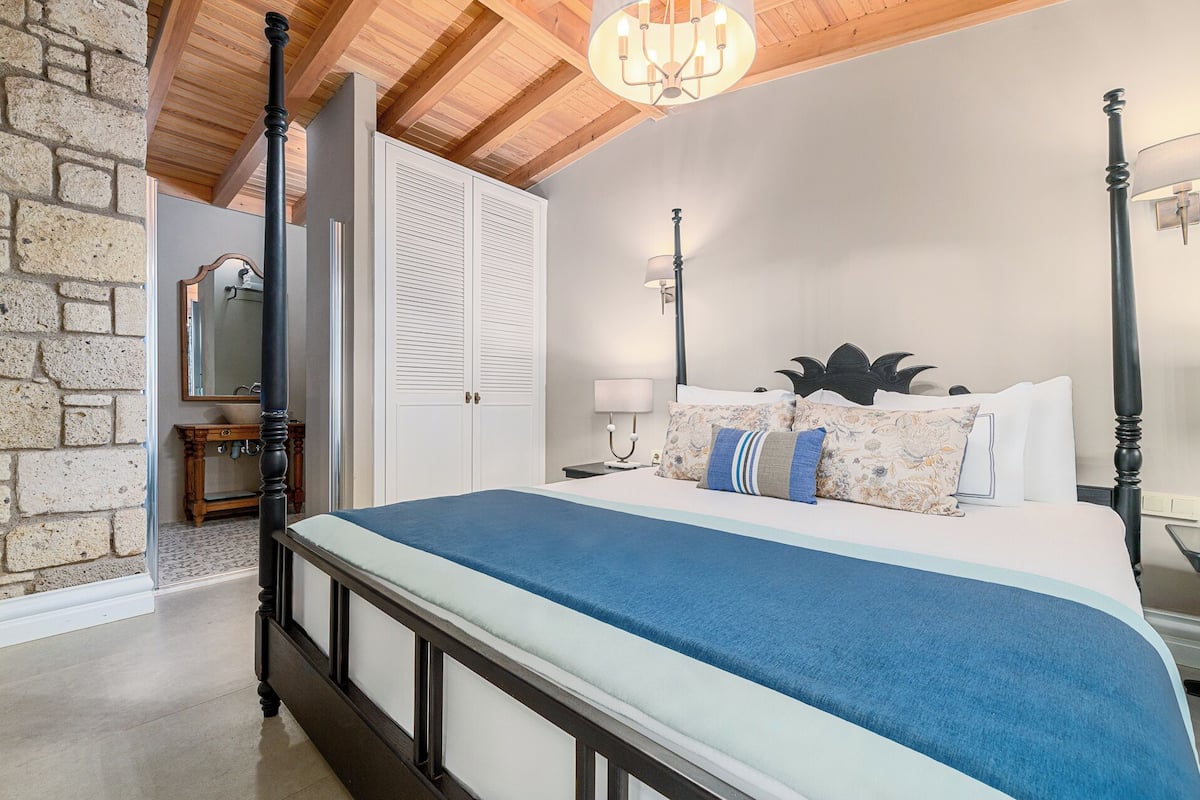 A bedroom with a four-poster bed, a stone wall, a wooden ceiling, and a chandelier.