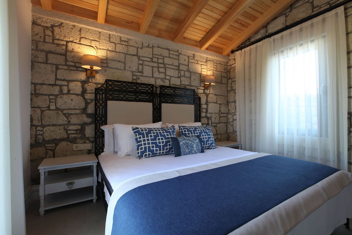 A bedroom with a stone wall, a wooden ceiling, a bed with a blue bedspread, two bedside tables with lamps, and a window with curtains.