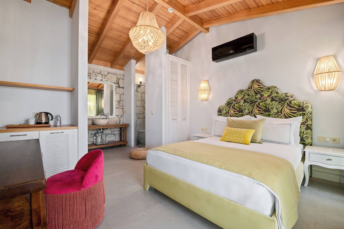 A bedroom with a bed, bedside tables, a desk, a chair, a shower, and a bathroom with a sink and mirror.