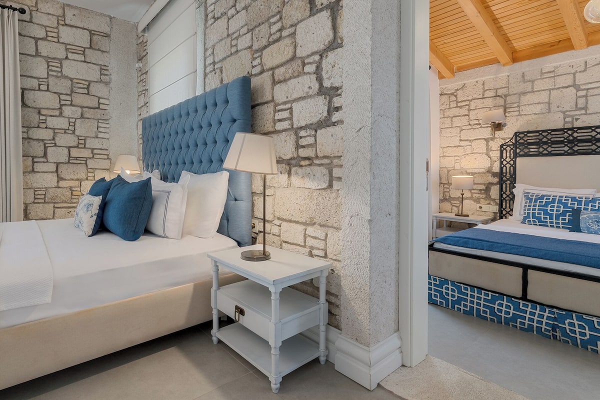 A bedroom with a stone wall, a bed with blue bedding, a bedside table with a lamp, and a nightstand with a lamp.