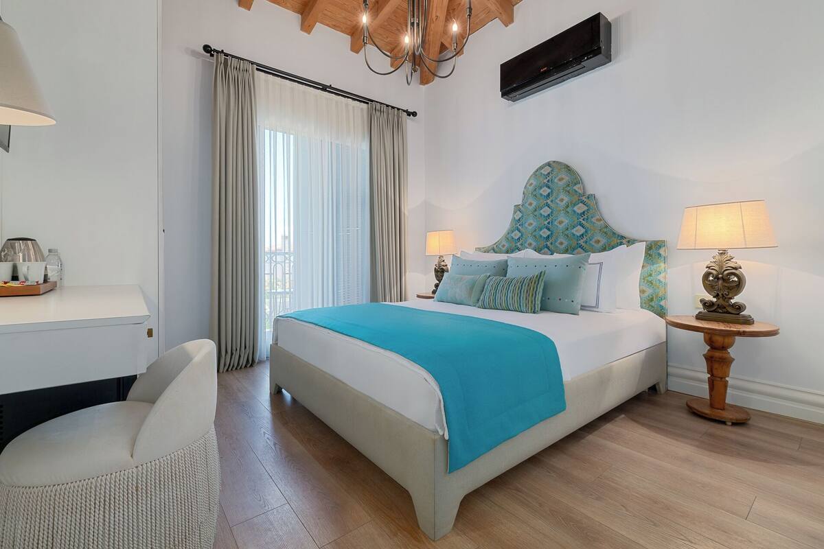 A bedroom with a bed, two bedside tables with lamps, a chair, a window with curtains, and a wooden ceiling.