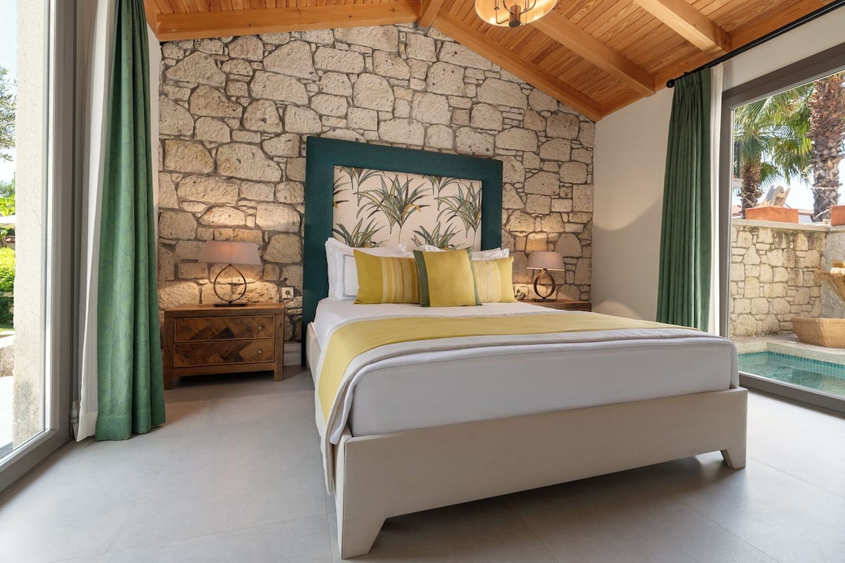 A bedroom with a stone wall, a bed with yellow and white bedding, two bedside tables with lamps, and a view of the outside.