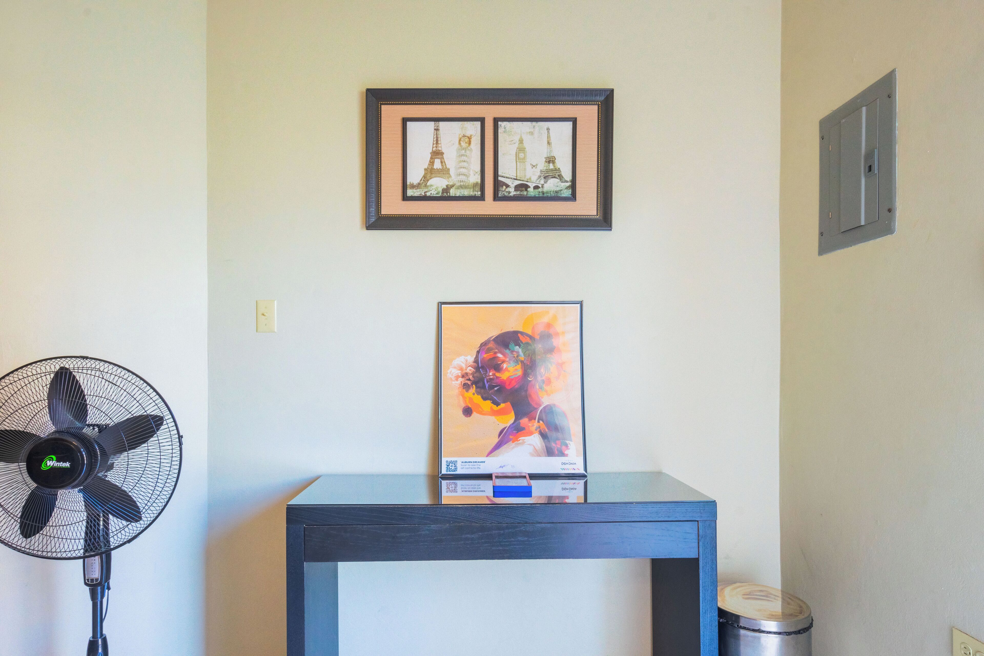 A room with a framed picture of the Eiffel Tower, a table with a framed picture, a standing fan, and a trash can.