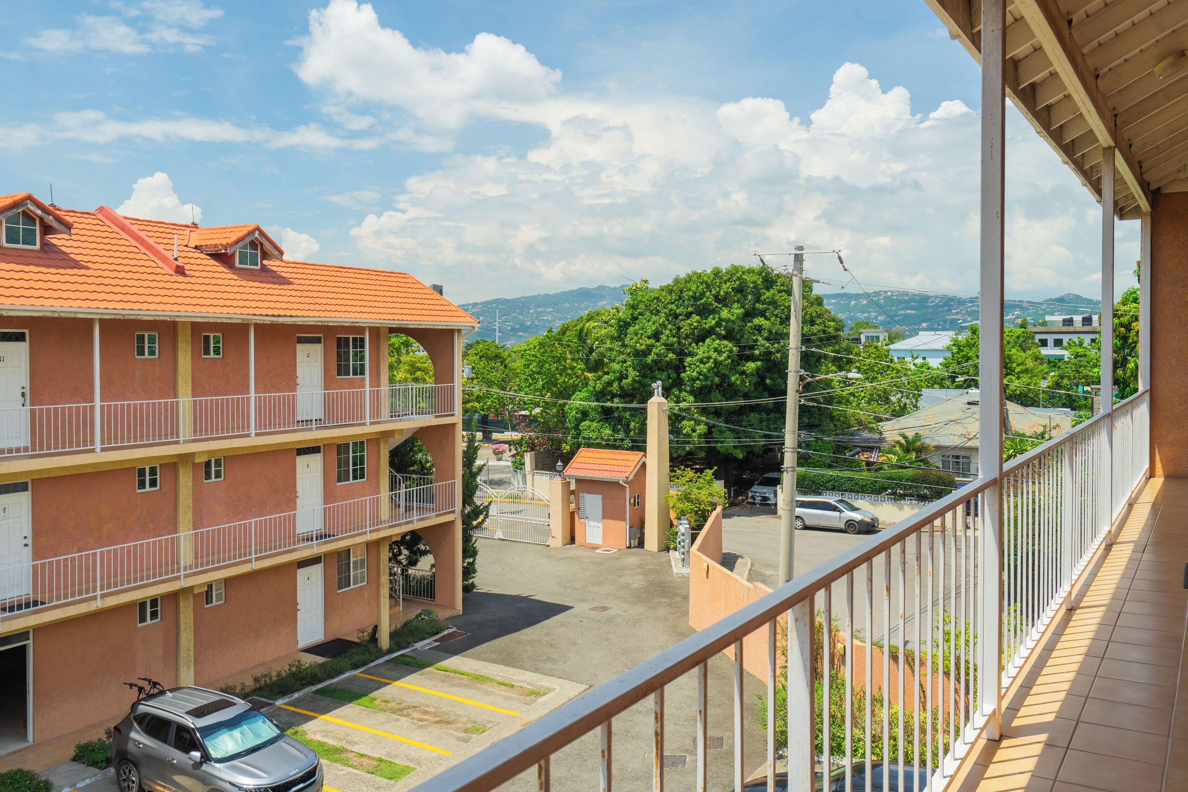 A multi-story building with balconies, a parking area, and a view of a green landscape and distant hills.