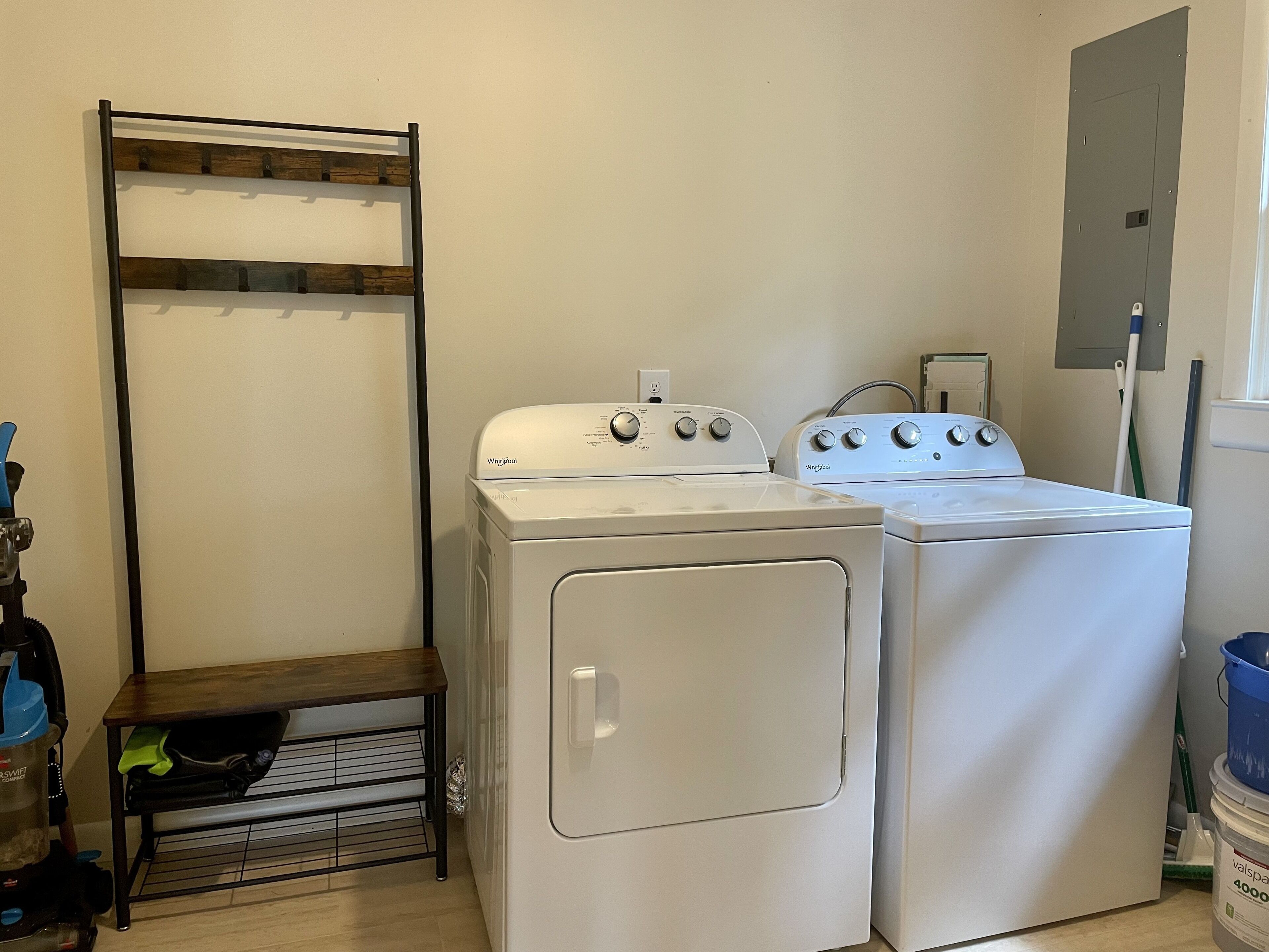 A laundry room with two white Whirlpool washers and dryers, a wooden shelf, and a metal drying rack.