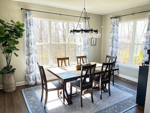 Un comedor con mesa y sillas de madera, ventanas amplias con cortinas, una lámpara colgante y una planta en maceta.