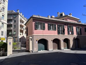 Un edificio rosa con contraventanas verdes, una entrada protegida por una reja y un cielo azul claro.