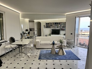 A modern, well-lit room with a dining area, a white sofa, and a balcony with a view.