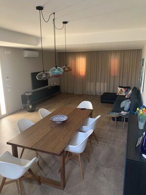 Ein modernes Wohnzimmer mit einem Holzessttisch, weißen Stühlen, einem dunklen Sofa und einem Fernseher auf einem Schrank.