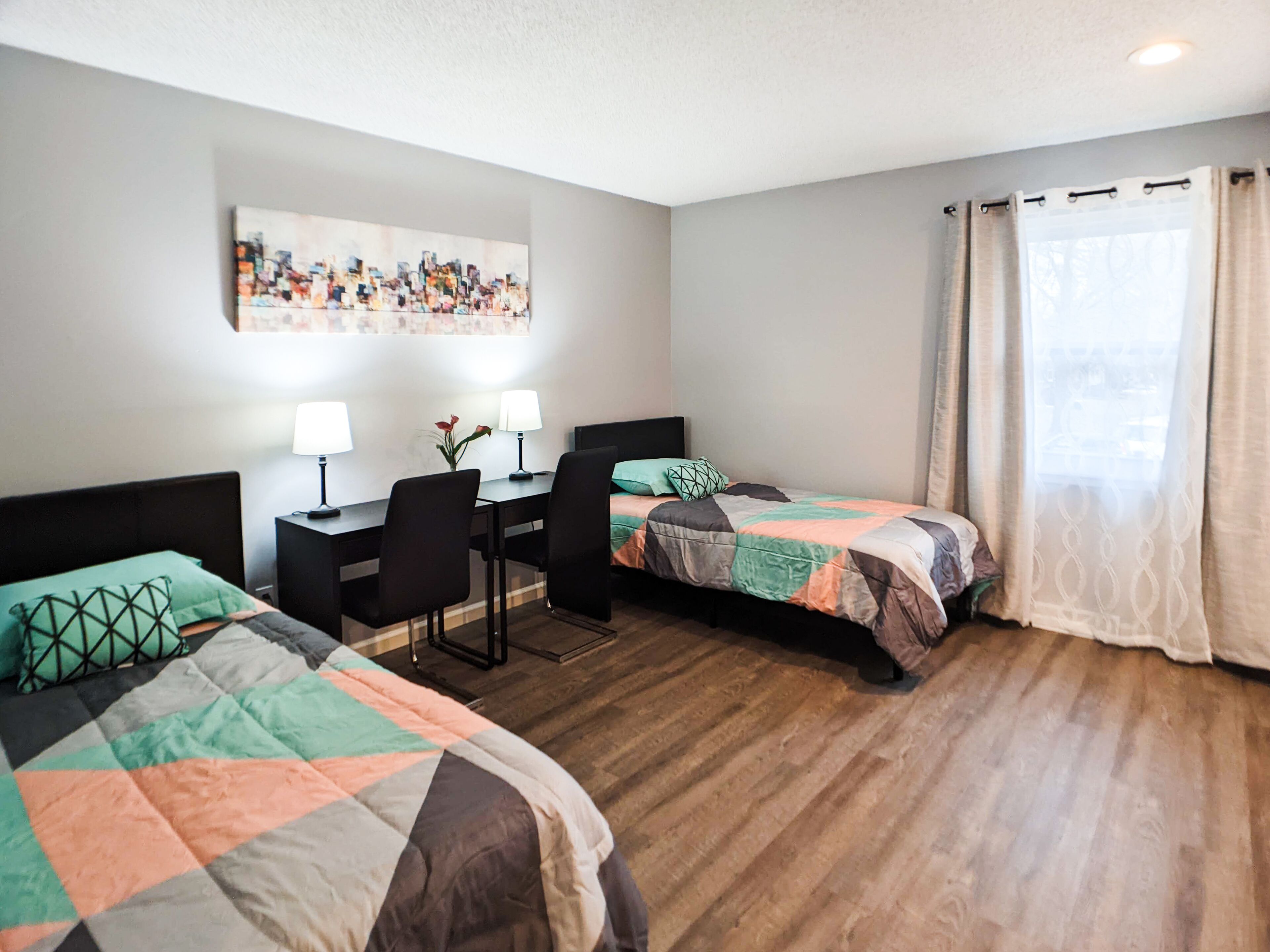 A hotel room with two beds, a desk with two lamps, a chair, and a painting on the wall.