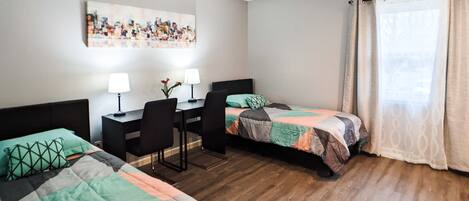 A hotel room with two beds, a desk with two lamps, a chair, and a painting on the wall.
