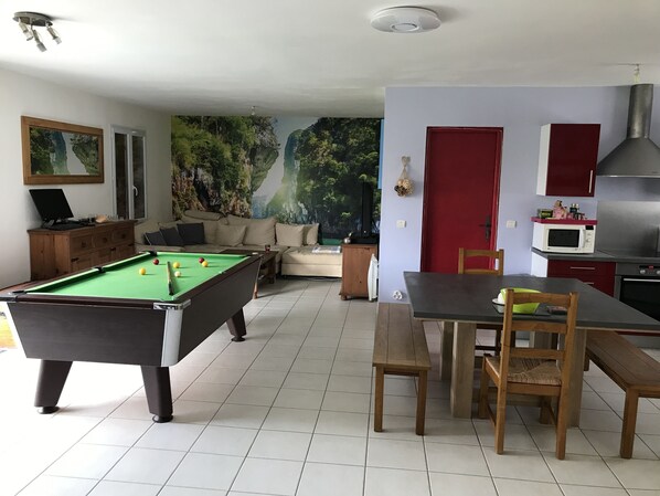 A living room with a pool table, a sofa, a dining table with chairs, a kitchen area with a microwave, and a red door.