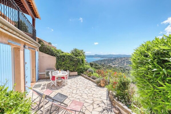 A patio with a table and chairs, a view of the sea, and a balcony with blue shutters.