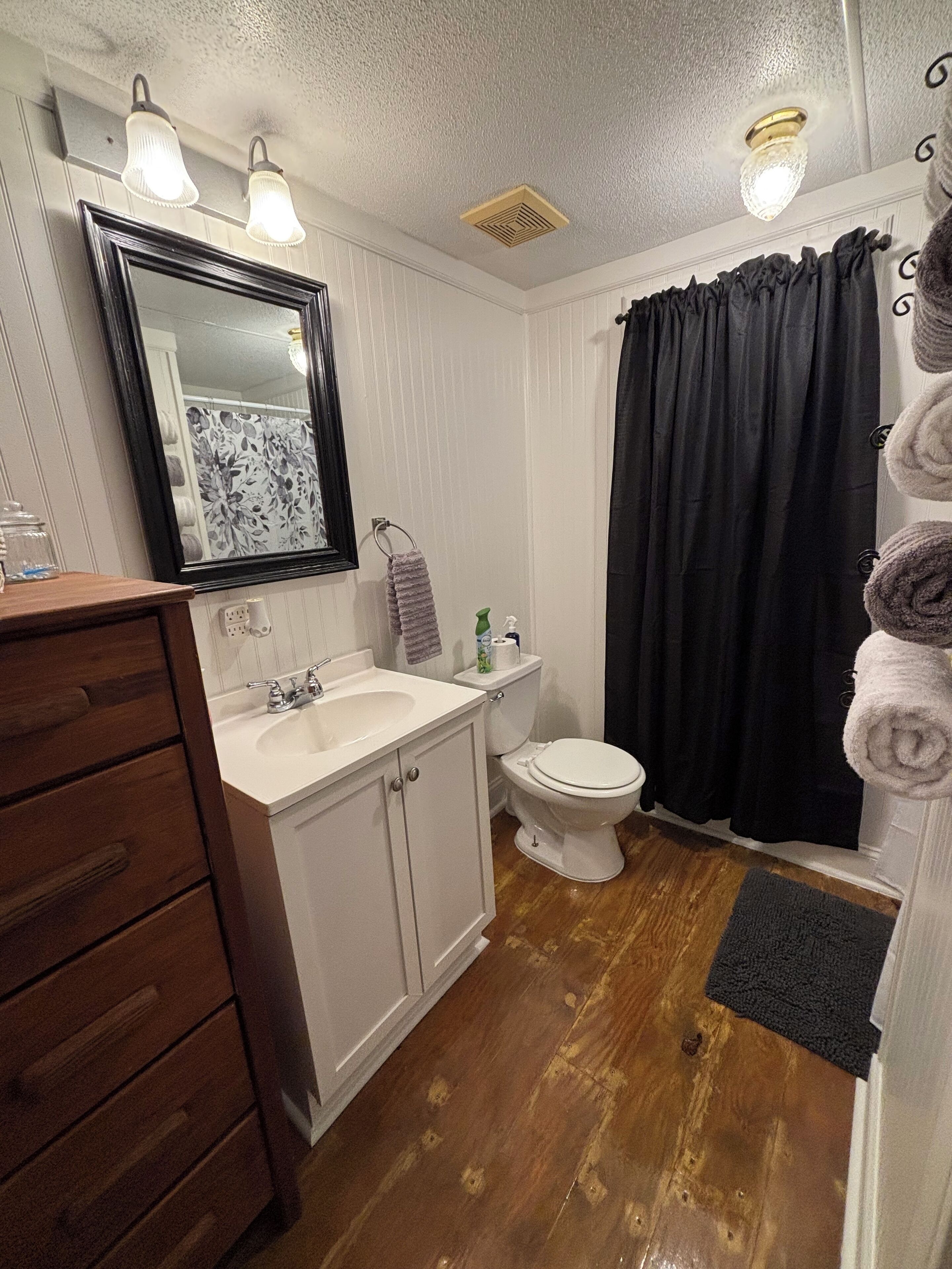 A bathroom with a wooden vanity, a white sink, a toilet, and a black shower curtain.
