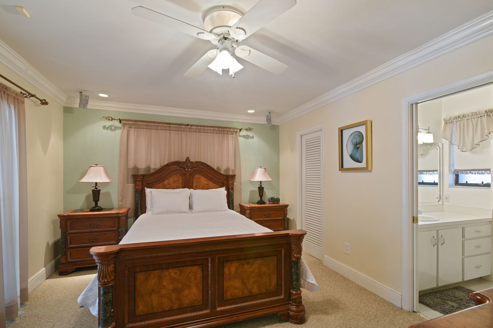 Une chambre à coucher avec un lit en bois, deux tables de chevet avec des lampes, un ventilateur de plafond, une armoire et une salle de bain avec un miroir et un lavabo.