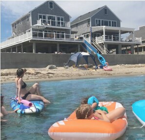 A beachfront property with a large house, a sandy beach, and inflatable rafts in the water. - Scituate oceanfront gem; 5BR  w/ wrap around deck. Perfect for family & friends (Scituate)