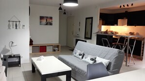Un salon moderne avec un canapé gris, une table basse blanche et une petite table d’appoint. On y trouve également un coin cuisine avec un bar et des tabourets.