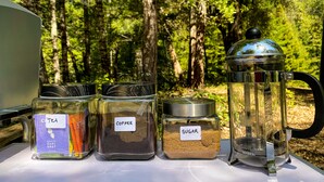 A table with labeled containers of tea, coffee, and sugar, and a French press coffee maker, set outdoors with a forest background. - Glamping in the Redwoods, with hot tub! The Knoll (Garberville)