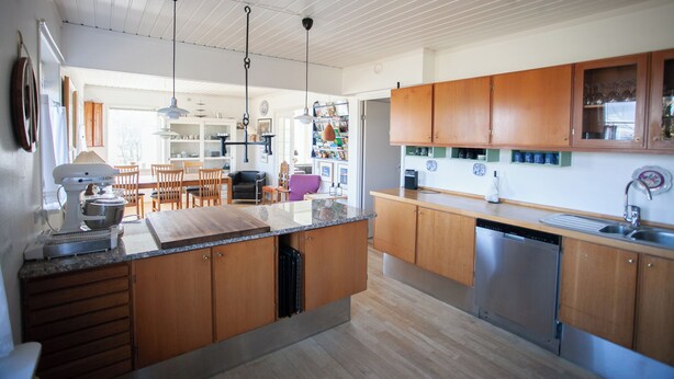 A modern kitchen with wooden cabinets, a central island, and a dishwasher. There is a dining area with chairs and a table visible in the background. - Lovely Apartments - 20 min walk from city centre (Tórshavn)