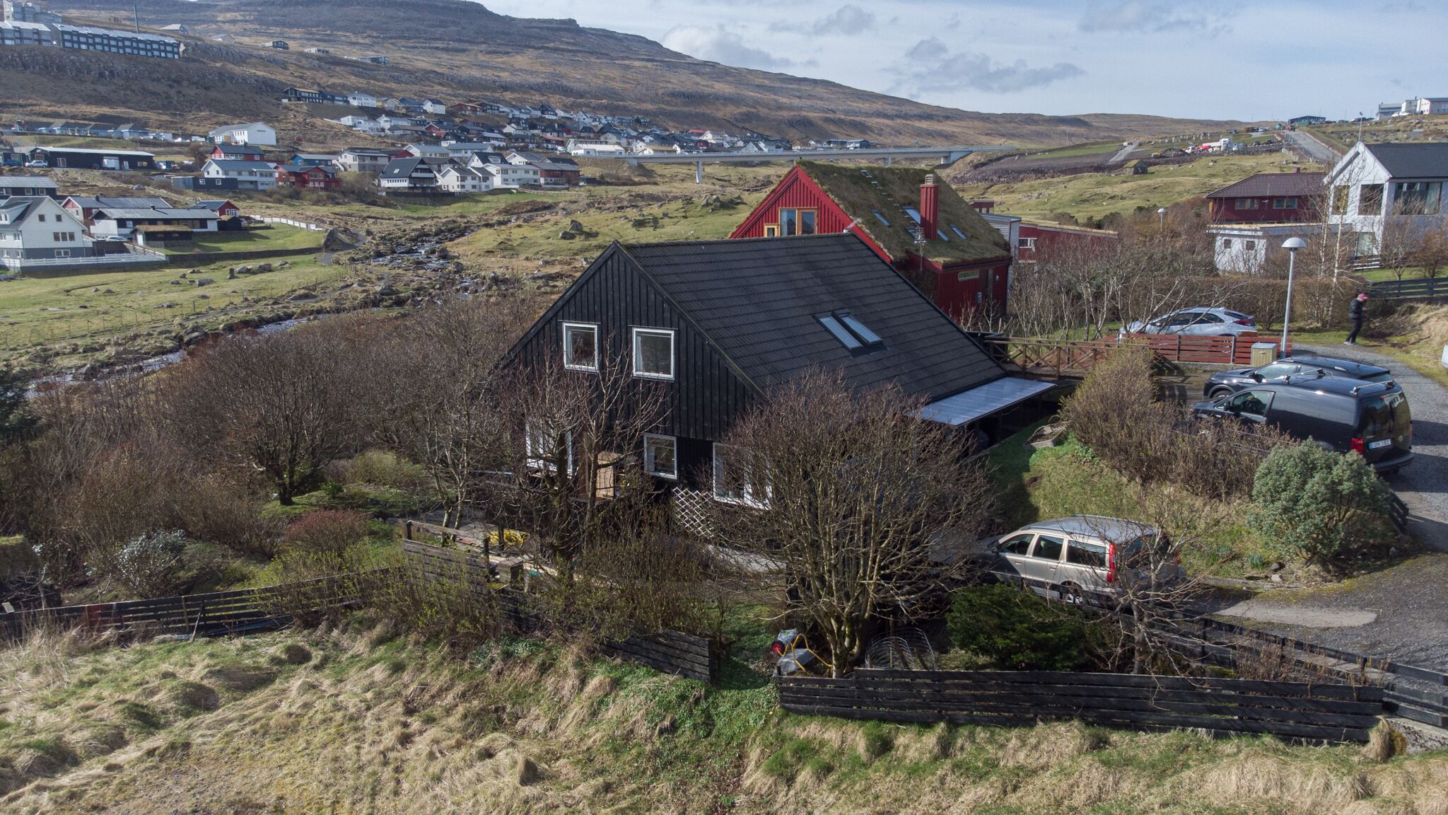 A rural house with a black roof and a red chimney, surrounded by a fenced yard and parked cars.